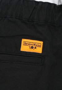 Mustast kangast tekstuuriga pind, millele on õmmeldud oranž silt tekstiga "Service Works" ning graafiliste ikoonidega.