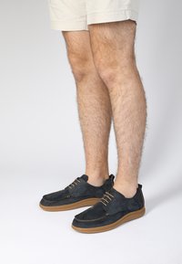 Dunkelblaue Wildleder-Schuhe mit braunen Sohlen, die ein minimalistisches Schnürdesign und kontrastierende Nähte aufweisen. Platziert mit nackten Beinen in Shorts.