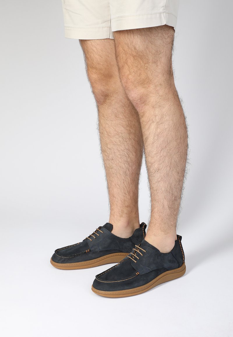 Dunkelblaue Wildleder-Schuhe mit braunen Sohlen, die ein minimalistisches Schnürdesign und kontrastierende Nähte aufweisen. Platziert mit nackten Beinen in Shorts.