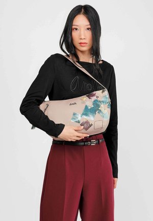 Bolso de hombro de cuero rosa con un diseño curvado, que presenta un collage de acentos de patrones azules y marrones; modelo vistiendo una camiseta negra y pantalones burdeos.