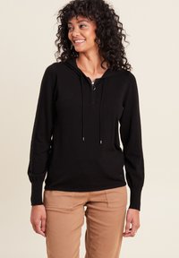 Schwarzer Kapuzenpullover mit einem halben Reißverschluss vorne, langen Ärmeln und Kordelzügen. Weiches Strickmaterial mit geripptem Saum. Getragen mit beigen Hosen.