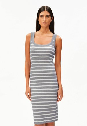 Vrouw staand in een mouwloze, grijze jurk met horizontale witte strepen, kijkend naar voren tegen een effen witte achtergrond.