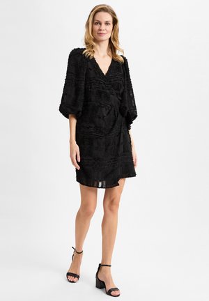 Schwarzes Kleid aus strukturiertem Stoff, mit V-Ausschnitt und weiten Ärmeln. Oberhalb des Knies geschnitten mit tailliertem Schnitt. Kombiniert mit schwarzen Ankle-Strap-Pumps.