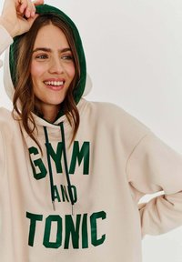 Béžová mikina s kapucí a zelenými šňůrkami, na přední straně s velkým zeleným nápisem "GYM AND TONIC." Má kontrastní kapuci se zeleným lemováním. Příjemně měkká textura.