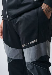 Pantalones deportivos negros con paneles perforados grises, un diseño elegante y el texto "PROJECT X PARIS" en la parte baja de la pierna.