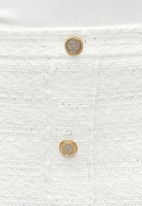 Λευκό ύφασμα με υφασμένες ρίγες και παγέτες· διαθέτει χρυσά κουμπιά διακοσμημένα με κυκλικούς στρας για κλείσιμο.