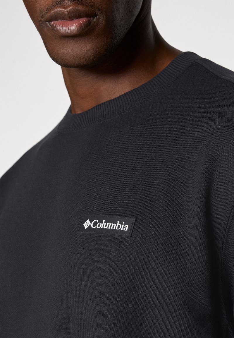 Sweatshirt noir en tissu tricoté avec un col ras du cou côtelé et un patch rectangulaire du logo Columbia sur la poitrine.