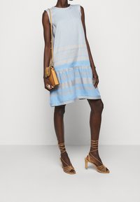 Hellblaues, ärmelloses Kleid mit strukturierten Mustern und geschichteter Gestaltung. Tanfarbene Riemchen-Absätze und eine kleine tanfarbene Tasche mit einer Kettenverzierung.