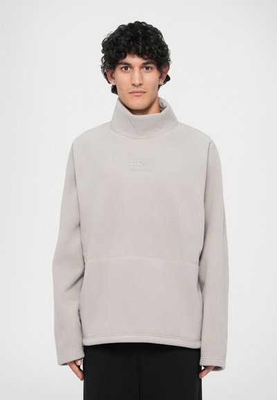 Pull léger gris clair avec un col haut, fabriqué en tissu polaire doux. Présente un logo embossé subtil et une poche kangourou.