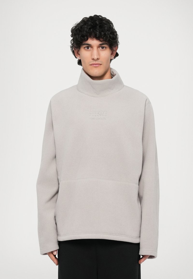 Světle šedý pulovr s vysokým límcem, vyrobený z měkkého fleece materiálu. Obsahuje jemně embosované logo a klokanní kapsu.