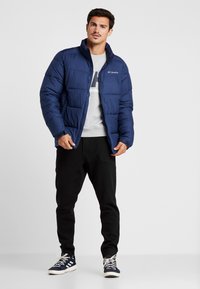 Chaqueta acolchada azul marino con diseño acolchado, cuello alto y logo de marca. Lleva puesta sobre un sudadera gris, con pantalones de ch ándal negros y zapatillas.