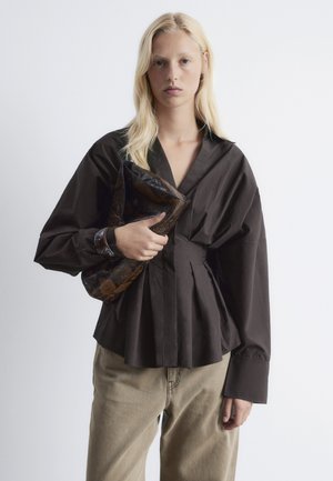 Dunkelbraune Bluse mit tiefem V-Ausschnitt, plissiertem Vorderteil und langen puffartigen Ärmeln. Kombiniert mit beigen Hosen und einer strukturierten Clutch.
