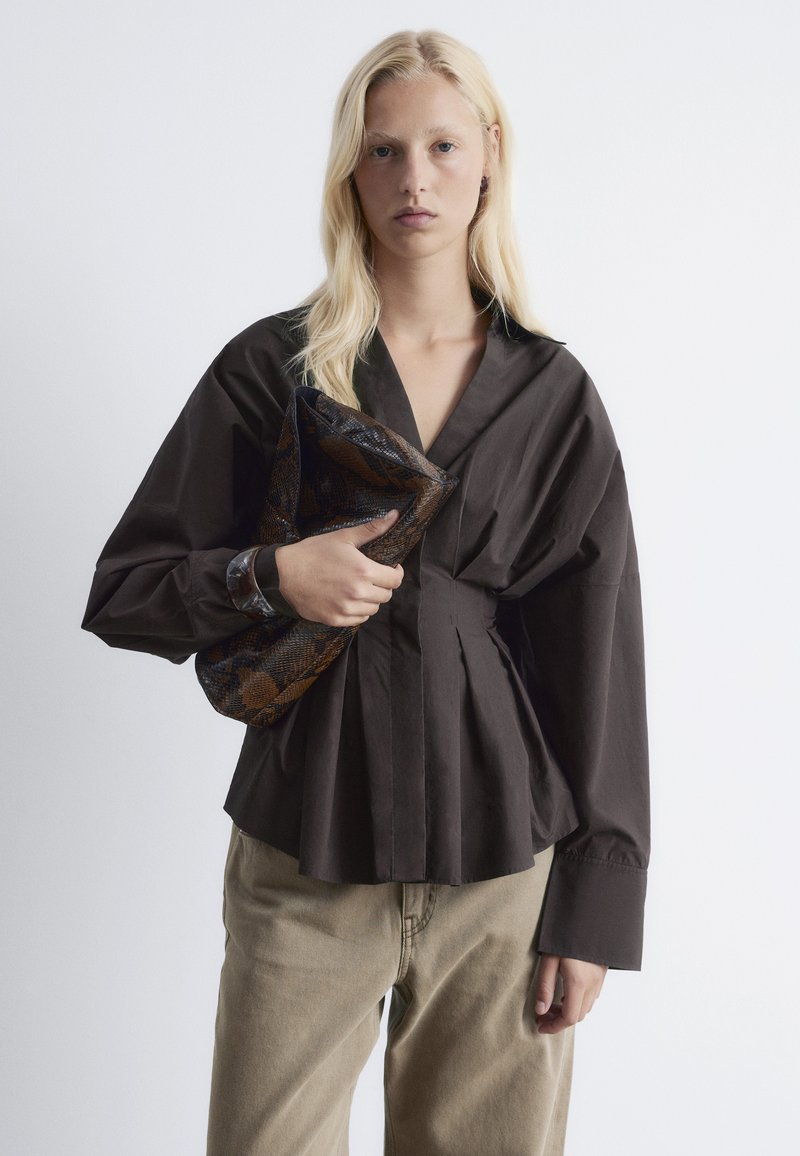 Dark brown blouse with a deep V-neck, pleated front, and long puff sleeves. Paired with beige pants and a textured clutch.