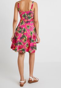 Robe d'été rose ajustée avec un imprimé de feuilles tropicales vertes et de fleurs, jupes évasée, larges bretelles et texture lisse. Portée avec des sandales blanches.