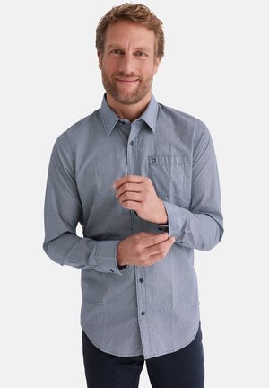 Mann lächelnd, der den Ärmelmanschetten seines hellblauen langärmligen Hemdes mit Muster und Brusttasche anpasst, vor einem schlichten weißen Hintergrund stehend.