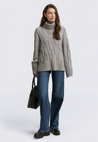 Debel siv pleteni pulover z vzorcem klic, visokim ovratnikom, robom z rebrastim vzorcem, kombiniran z razširjenimi modrimi kavbojkami in črnimi platformnimi škornji, drži črno torbo.