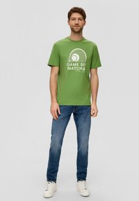 T-shirt di cotone verde con grafica di tennis, maniche corte e collo rotondo. Abbinato a jeans in denim blu e sneakers bianche.