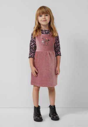 Robe en velours côtelé rose avec bretelles, ornée de broderies florales ; associée à une chemise noire à manches longues avec motifs floraux et des bottes noires à lacets.