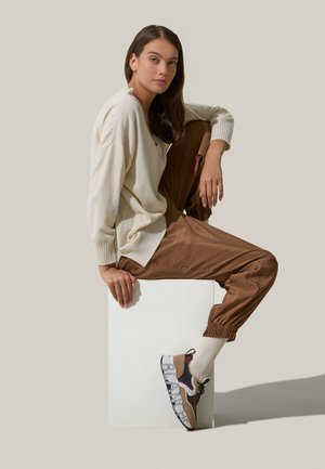 Maglione bianco a maglia con polsini a coste, pantaloni beige in vita alta e scarpe da ginnastica con dettagli a blocchi di colore e materiali testurizzati.