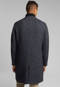 Uomo che indossa un cappotto di lana a spina di pesce grigio scuro con colletto nero, in piedi di spalle contro uno sfondo grigio uniforme.
