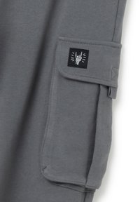 Sudo de color gris con una bolsa lateral que presenta un parche negro y un símbolo de gesto de mano bordado en blanco. Textura suave, diseño casual.