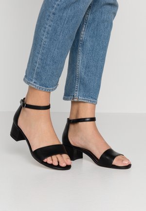 Sandales à talons en cuir noir avec un design à deux brides et boucle de cheville. Caractérisées par une pointe carrée et un petit talon carré. Portées avec un jean bleu raccourci.