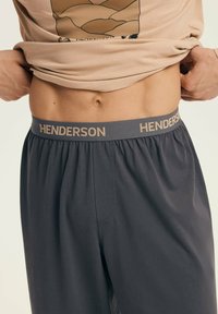 Midsection of person lifting beige shirt, wearing dark gray pants with elastic waistband labeled "HENDERSON."
