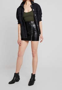 Veste en denim noire sur un débardeur vert olive, associée à une mini-jupe en cuir noire avec une fermeture éclair avant. Des bottines noires à la cheville complètent le look.