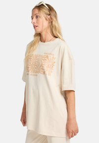 Beige oversized t-shirt lavet af bomuld, med en blomster grafik i kobberfarver på forsiden og korte ærmer.