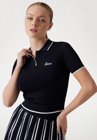 Marineblaues Polohemd mit kurzen Ärmeln, gerippter Textur, weißem Saum und gesticktem "Guess"-Logo auf der Brust, kombiniert mit einem schwarz-weißen gestreiften Rock.