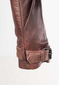 Mouw van een bruine leren jas met een gestructureerde afwerking, voorzien van een gespsluiting dichtbij de manchette en zichtbare stiksels langs de naad.