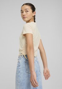 Camiseta de manga corta beige con detalle fruncido a los lados, combinada con una falda de mezclilla azul claro con bolsillos visibles y un ajuste relajado.