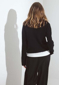 Melna adīta džemperis ar brīvu piegriezumu, uzvilkts virs baltās krekla. Modelis ir vērsts prom, ar viļņainiem matiem un ēnu, kas krīt uz gaišās sienas.