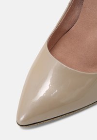Beżowe lakierowane czółenka z wydłużonym noskiem o gładkim, błyszczącym wykończeniu, odsłaniające miękkie zamszowe wnętrze oraz jasną, fakturowaną podeszwę.