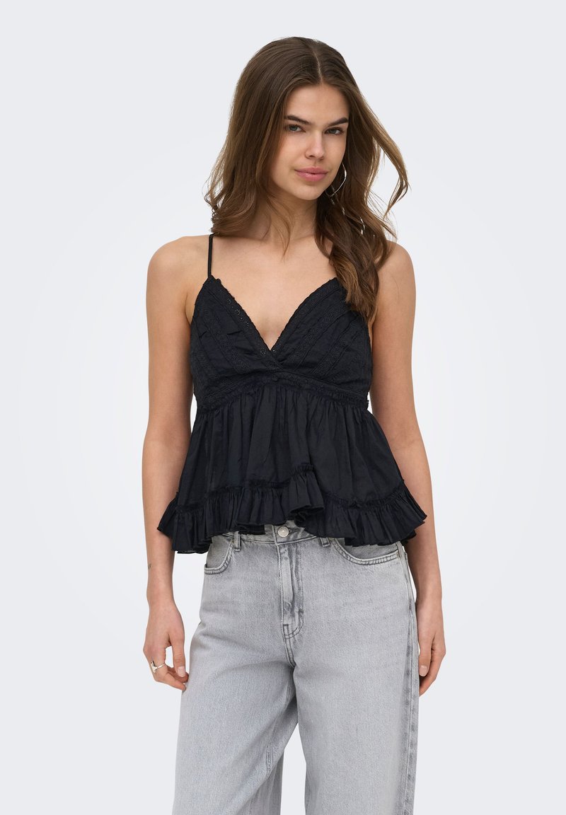 Jeune femme aux longs cheveux bruns portant un top noir sans manches à volants et un jean gris clair taille haute sur un fond uni.
