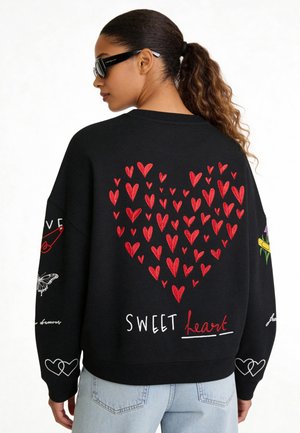 Donna con coda di cavallo che indossa una felpa nera con un cuore rosso composto da cuori più piccoli e le parole "SWEET heart" sulla schiena, jeans azzurri.