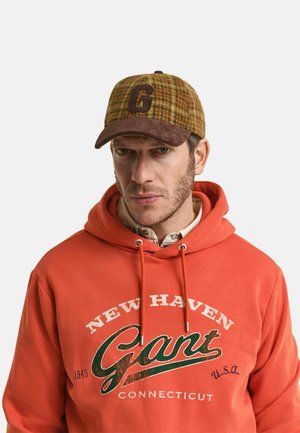 Orangener Hoodie mit dem Schriftzug "NEW HAVEN Gant U.S.A.". Brauner Karo-Hut mit einem großen "G" auf der Vorderseite. Lässiger, geschichteter Kleidungsstil.
