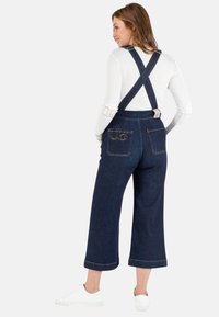 Dunkelblaue Denim-Latzhose mit weitem Bein, verstellbaren Trägern und bestickten Gesäßtaschen, kombiniert mit einem enganliegenden weißen Langarmoberteil und weißen Sneakers.