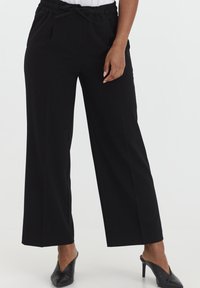 Zwarte wijde broek gemaakt van een gladde stof. Heeft een elastische tailleband met een strikdetail en zijzakken. Gecombineerd met zwarte muiltjes.