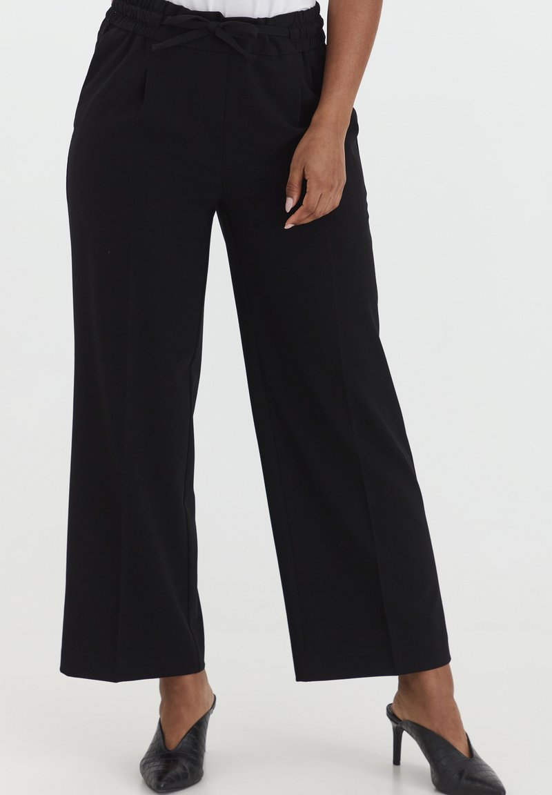 Zwarte wijde broek gemaakt van een gladde stof. Heeft een elastische tailleband met een strikdetail en zijzakken. Gecombineerd met zwarte muiltjes.
