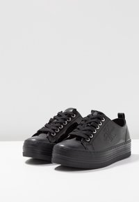 Black patent sneaker with rounded toe, thick striped sole, and black laces. Features logo detail on the side and a pull tab at the heel.