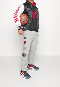 Chaqueta varsity de satén negra con acentos rojos y blancos, pantalones de chándal grises con gráficos y zapatillas deportivas coloridas. Sosteniendo un balón de baloncesto.