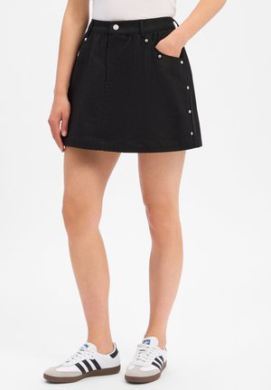 Femme portant une mini-jupe noire avec des boutons argentés sur le côté, un haut blanc et des baskets blanches avec des bandes noires sur fond blanc.