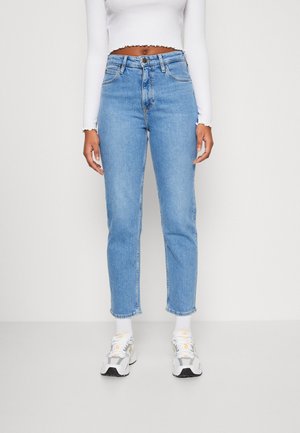 Femme portant un jean taille haute bleu clair droit, un haut blanc court à manches longues, des chaussettes blanches et des baskets blanches sur un fond uni.