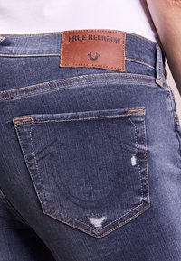 Tmavě modré džíny z denimu s opotřebovanými detaily, oranžovým prošitím a hnědou koženou nášivkou označenou "True Religion" nad zadní kapsou.