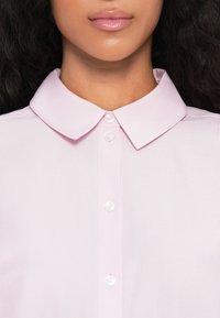 Chemise col boutonné rose clair avec des côtes verticales, dotée d'un plastron de boutons et de boutons brillants, mettant en avant une texture de tissu lisse.