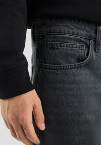 Graue Jeans aus Denim mit einer strukturierten Oberfläche, detaillierten Nähten und einer traditionellen Tasche. Kombiniert mit einem schwarzen Sweatshirt mit gerippten Bündchen.