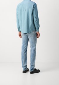 Camicia a maniche lunghe blu chiaro con polsini a bottone, abbinata a jeans chiari e scarpe nere, mostrata da dietro.