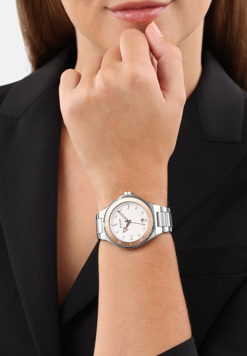 Silberner Metallarmbanduhr mit einem runden weißen und roségoldenen Zifferblatt, ausgestattet mit einer strukturierten Lünette und einem verlinkten Armband, getragen am Handgelenk vor einem dunklen Hintergrund.