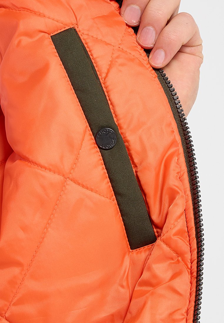 Main tenant la poche de la veste matelassée orange ouverte avec une fermeture éclair noire et un bouton-pression noir.